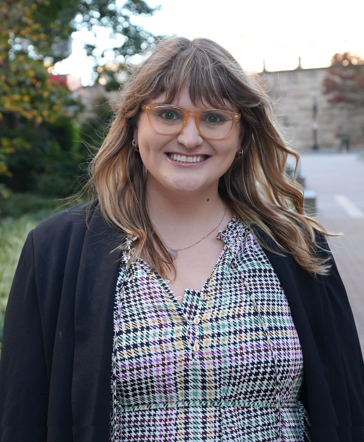 A woman with long brunette wavy hair, bangs and yellow glasses smiles at the camera. She is standing outdoors in front of greenery and a paved walkway. She is wearing a patterned dress and a black blazer.